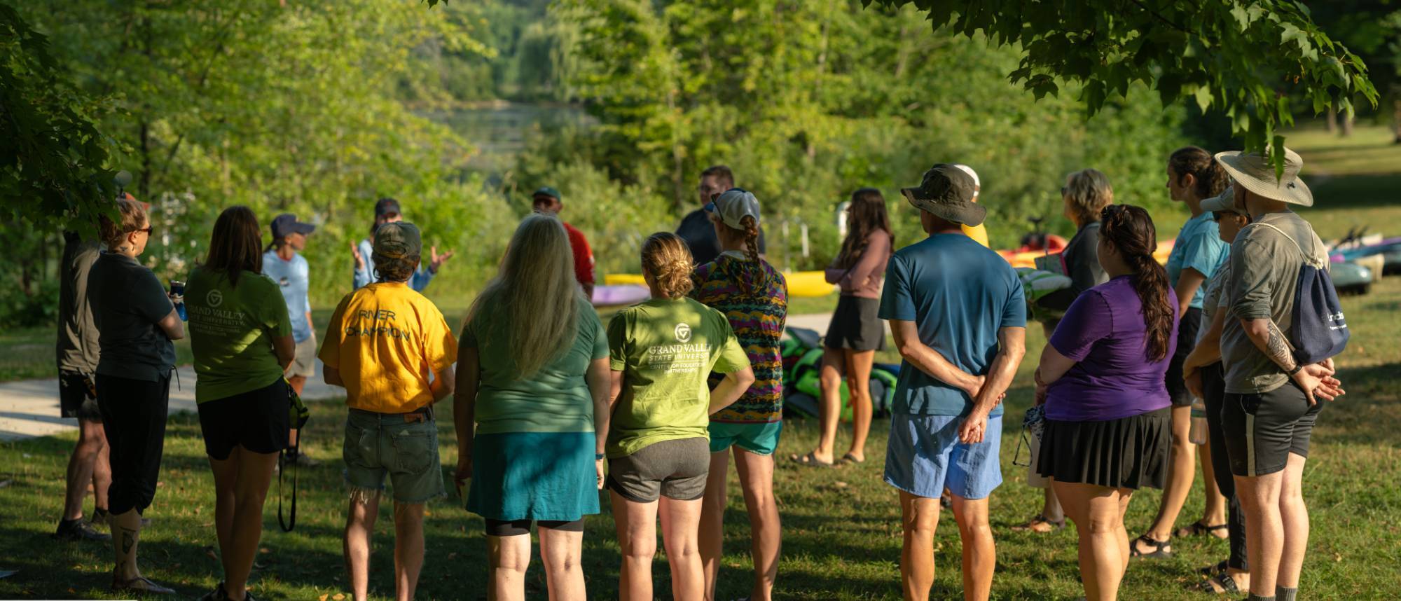 Adults in summer attire stand around in a circle listening to a speaker. About 20 people standing in the shade on a sunny day in the park.